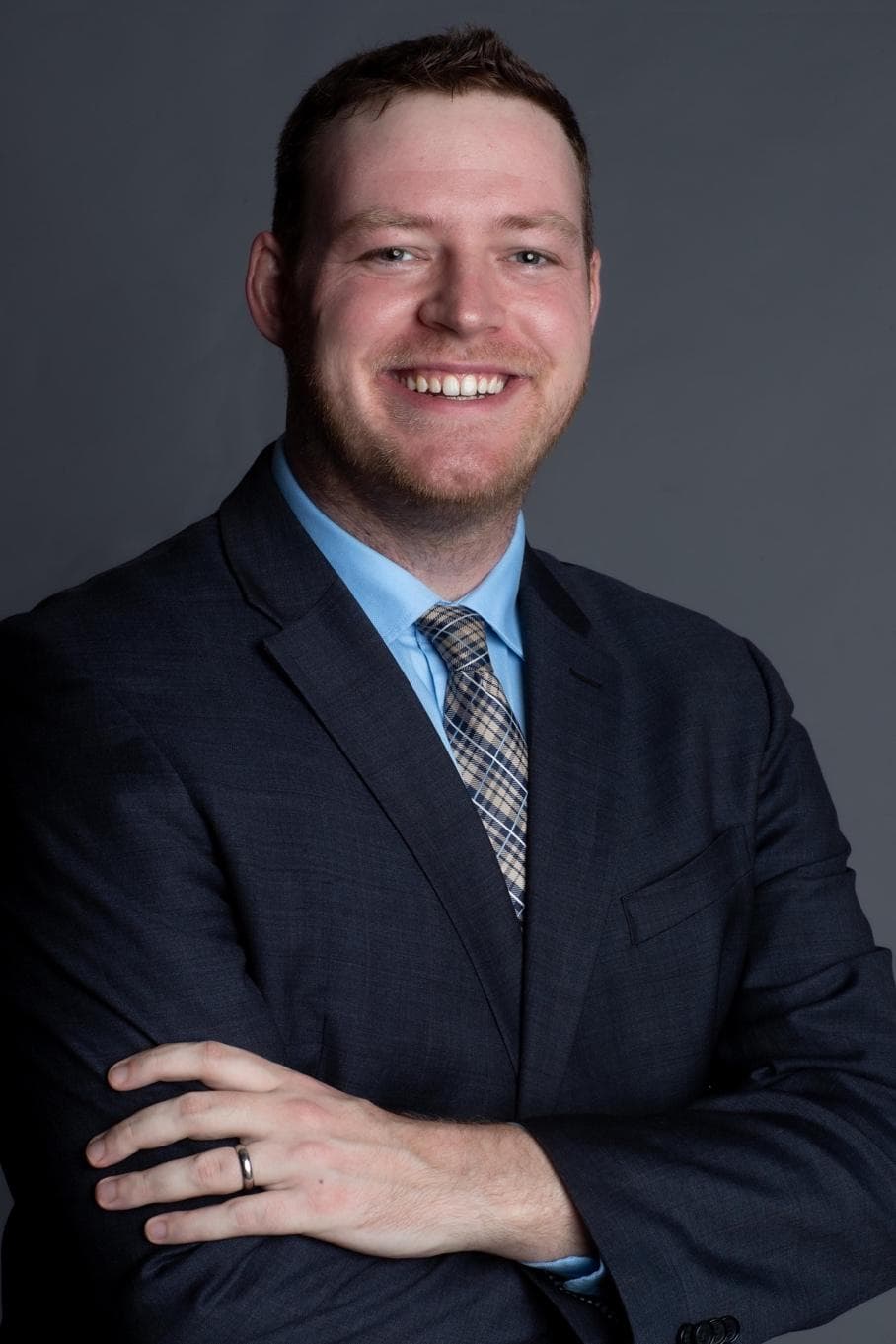 Smiling man in a dark suit and blue shirt with arms crossed against gray background.